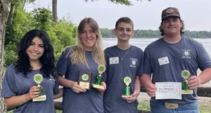 Four teens in grey t-shirts holding their 1st place trophies pose for a picture with Lake James in the background.